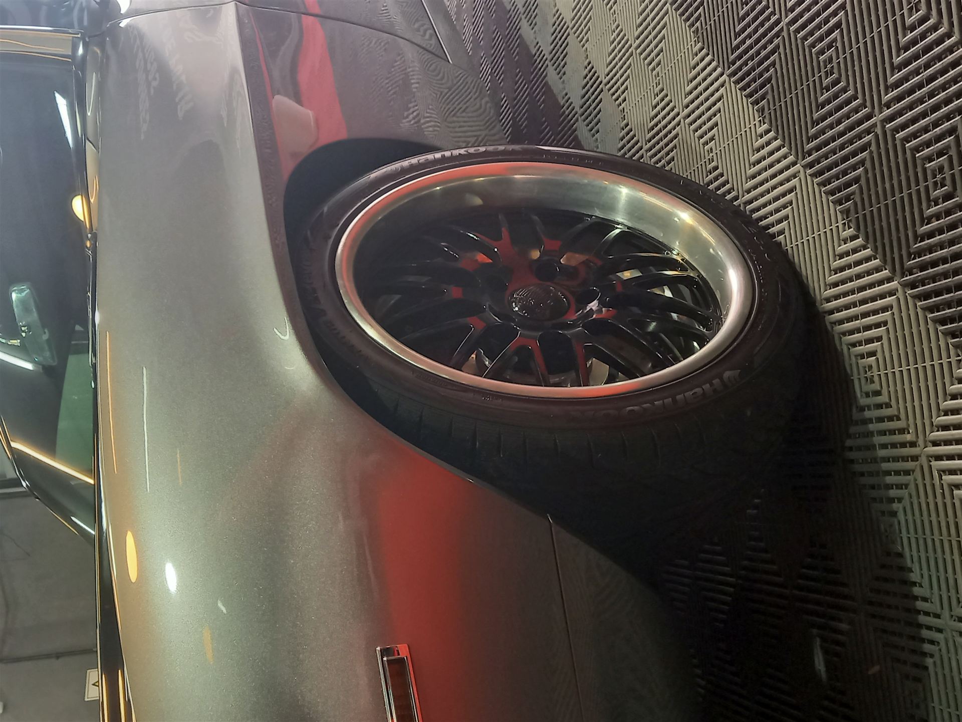 Detalle de llanta deportiva negra y roja con neumático Hankook, junto a un paragolpes trasero de alto brillo.
